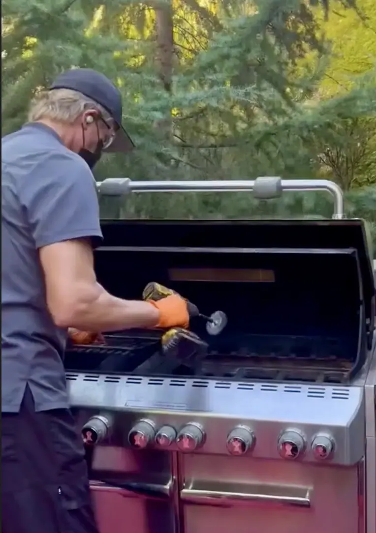 Greg from Grill Docs, wearing orange gloves, cleaning inside an open stainless gas grill with a drill-mounted brush.